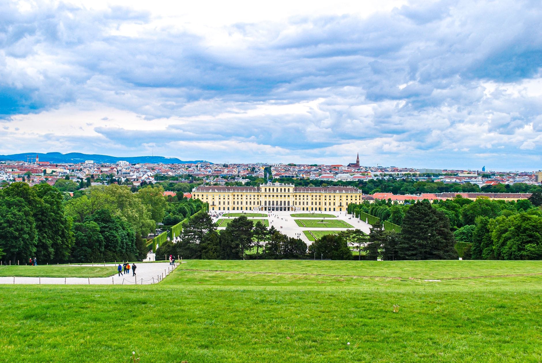 Weitwinkelansicht des Schönbrunner Schlosses mit Gärten und der Wiener Stadtkulisse im Hintergrund unter einem bewölkten Himmel.