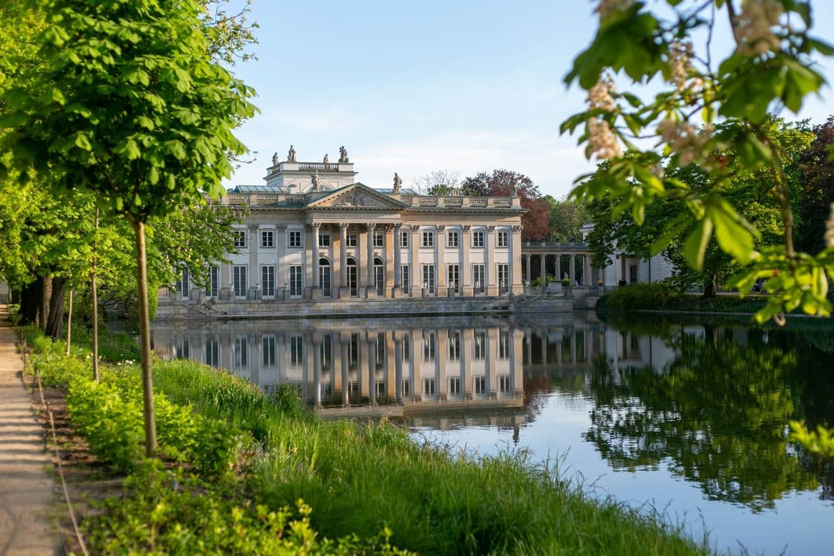 Vista del Palazzo Łazienki riflesso nell'acqua, circondato da lussureggianti alberi verdi e prato in una giornata di sole a Varsavia.