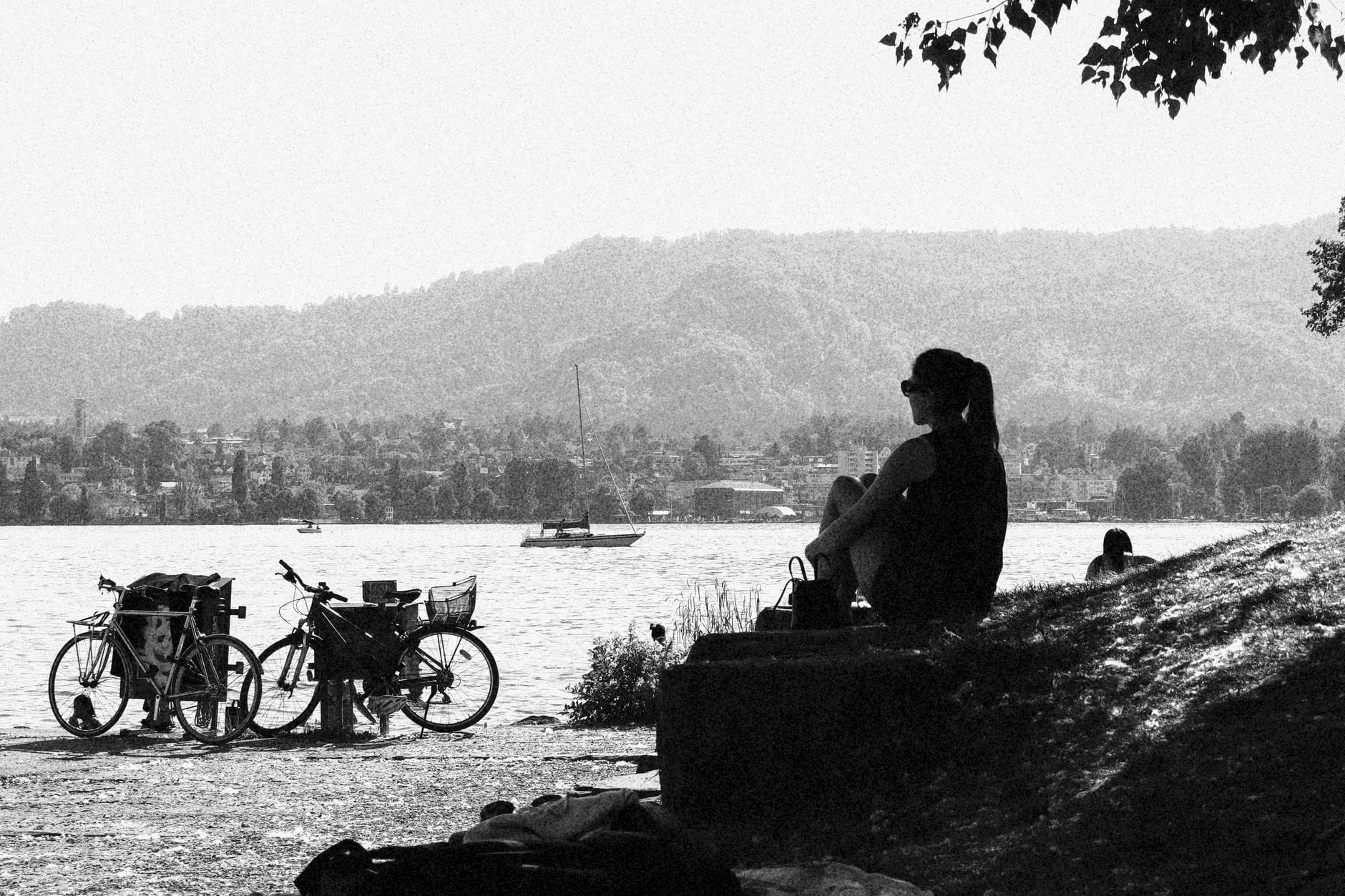 ภาพขาวดำของบุคคลนั่งอยู่บนฝั่งหญ้าริมทะเลสาบ Zürich มองเห็นจักรยานและเรือ มีต้นไม้และเนินเขาเป็นฉากหลัง