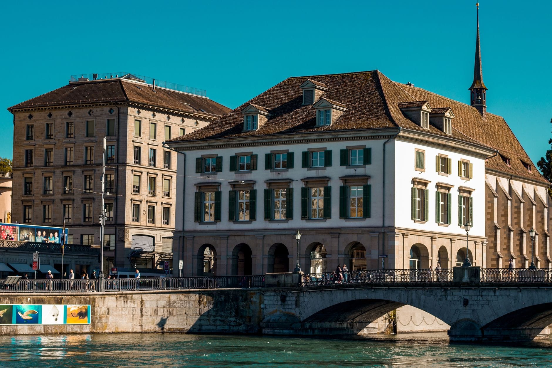 อาคารริมแม่น้ำสไตล์คลาสสิกใน Zürich พร้อมหน้าต่างโค้งและบานเกล็ดสีเขียว มองเห็นจากฝั่งตรงข้ามของแม่น้ำ วันที่แดดสดใสและมีผู้คนเดินอยู่ใกล้เคียง