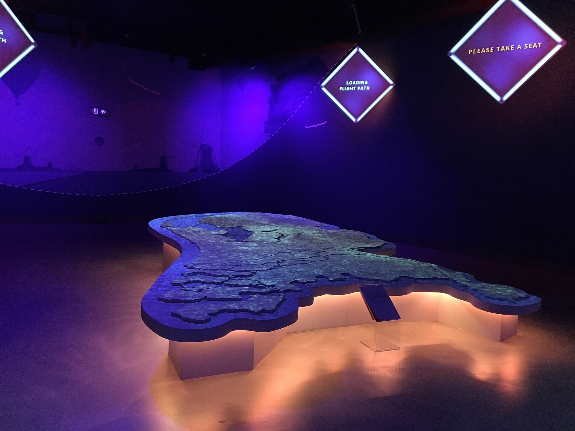 Un gran mapa 3D iluminado de los Países Bajos en el interior de THIS IS HOLLAND, con iluminación ambiental púrpura y letreros luminosos de rutas de vuelo en la pared.