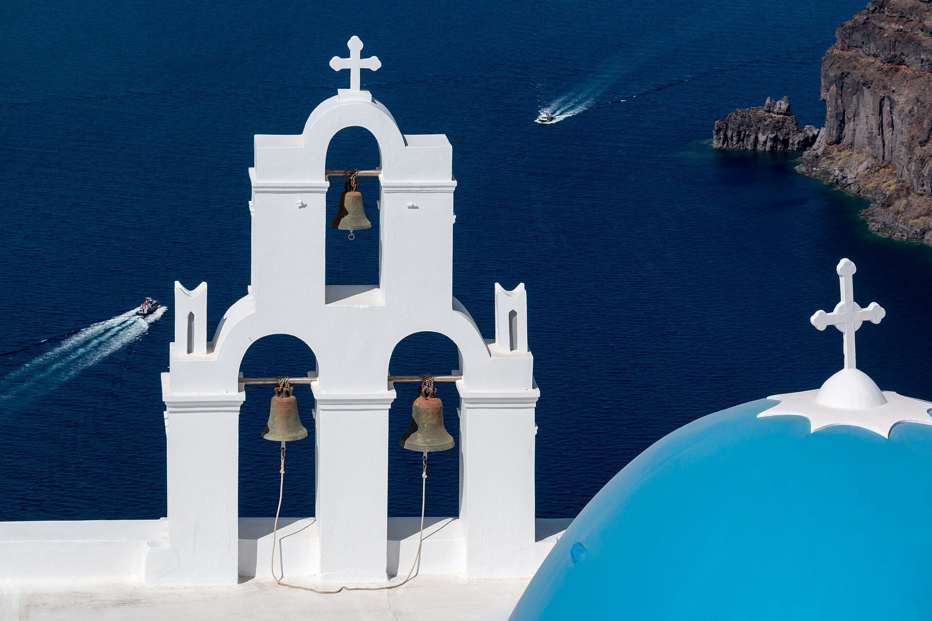 ระฆังสามใบของโบสถ์ Fira พร้อมหอระฆังสีขาว โดมสีน้ำเงิน และทะเล Aegean เป็นฉากหลัง