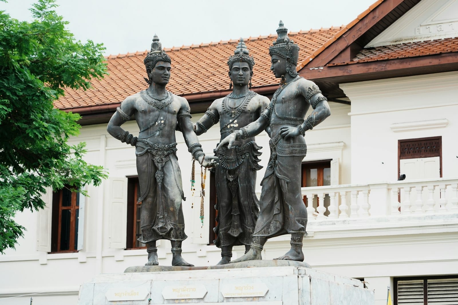 El Monumento a los Tres Reyes en Chiang Mai presenta tres estatuas de bronce sobre un pedestal de mármol frente a un edificio blanco con techo de tejas.