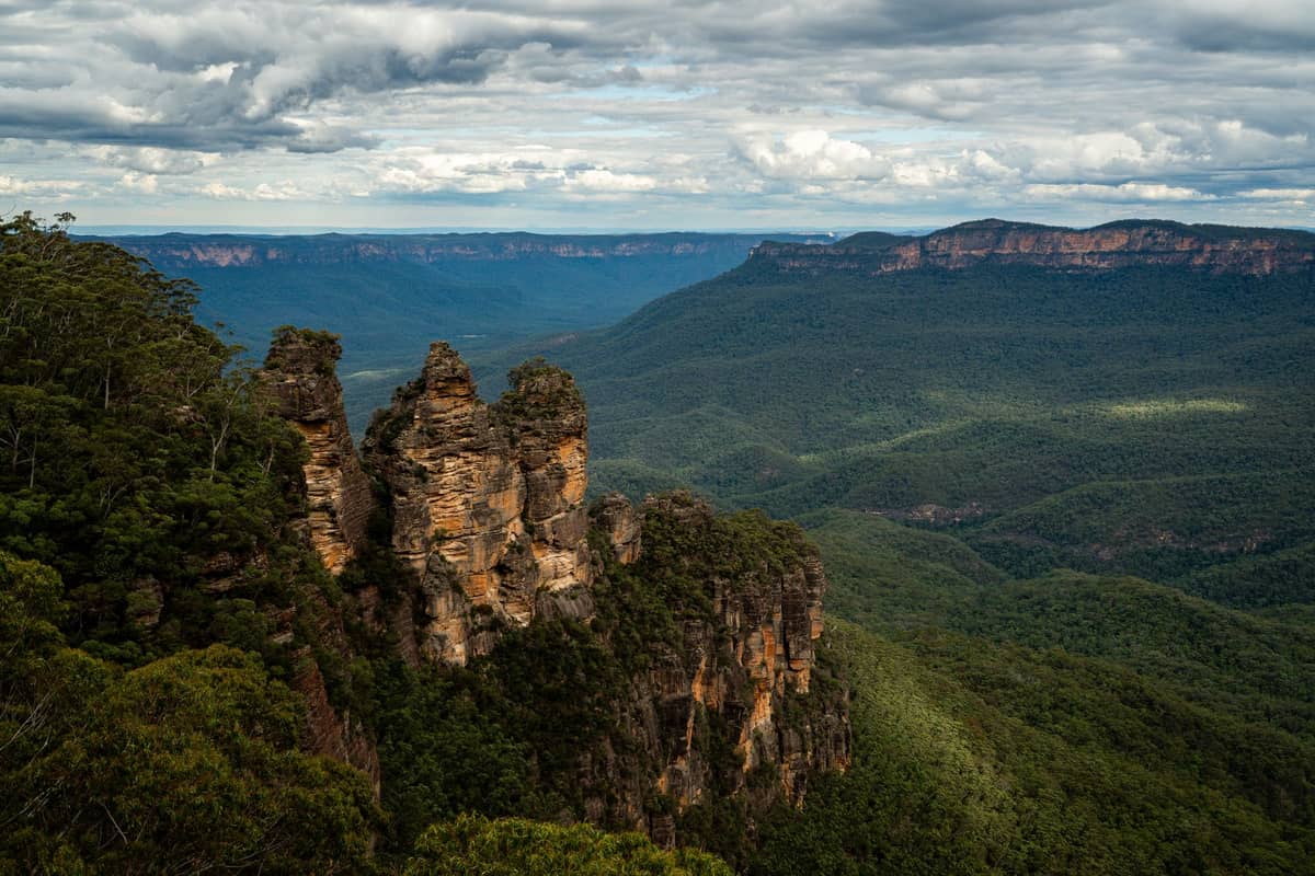 Wide view of the Three Sisters rock formation rising above the Jamison Valley, with dramatic clouds and expanses of forest, as seen from Echo Point in the Blue Mountains.