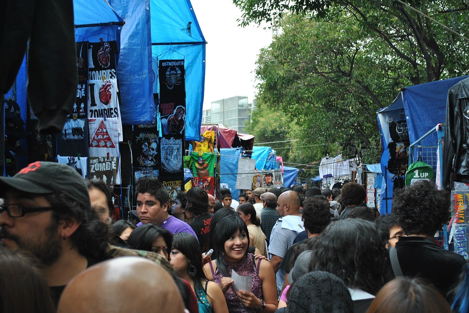 Le folle riempiono uno stretto vicolo fiancheggiato da bancarelle con teli blu e merce alternativa al Tianguis Cultural di Guadalajara, sotto alberi rigogliosi e verdi.