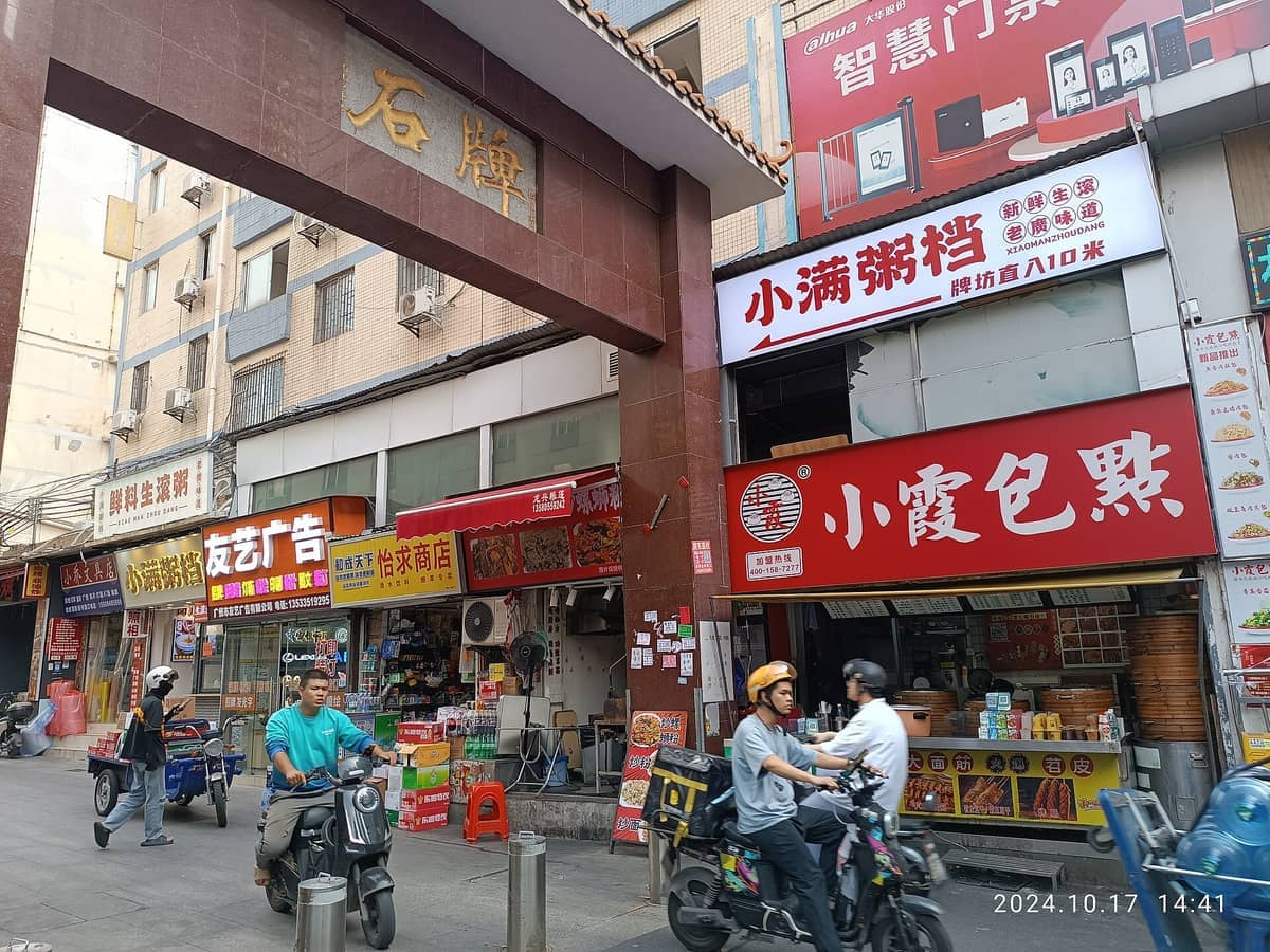 Street view of Tianhe in Guangzhou, featuring local eateries, colorful shop signs, and people riding scooters in a lively urban setting.