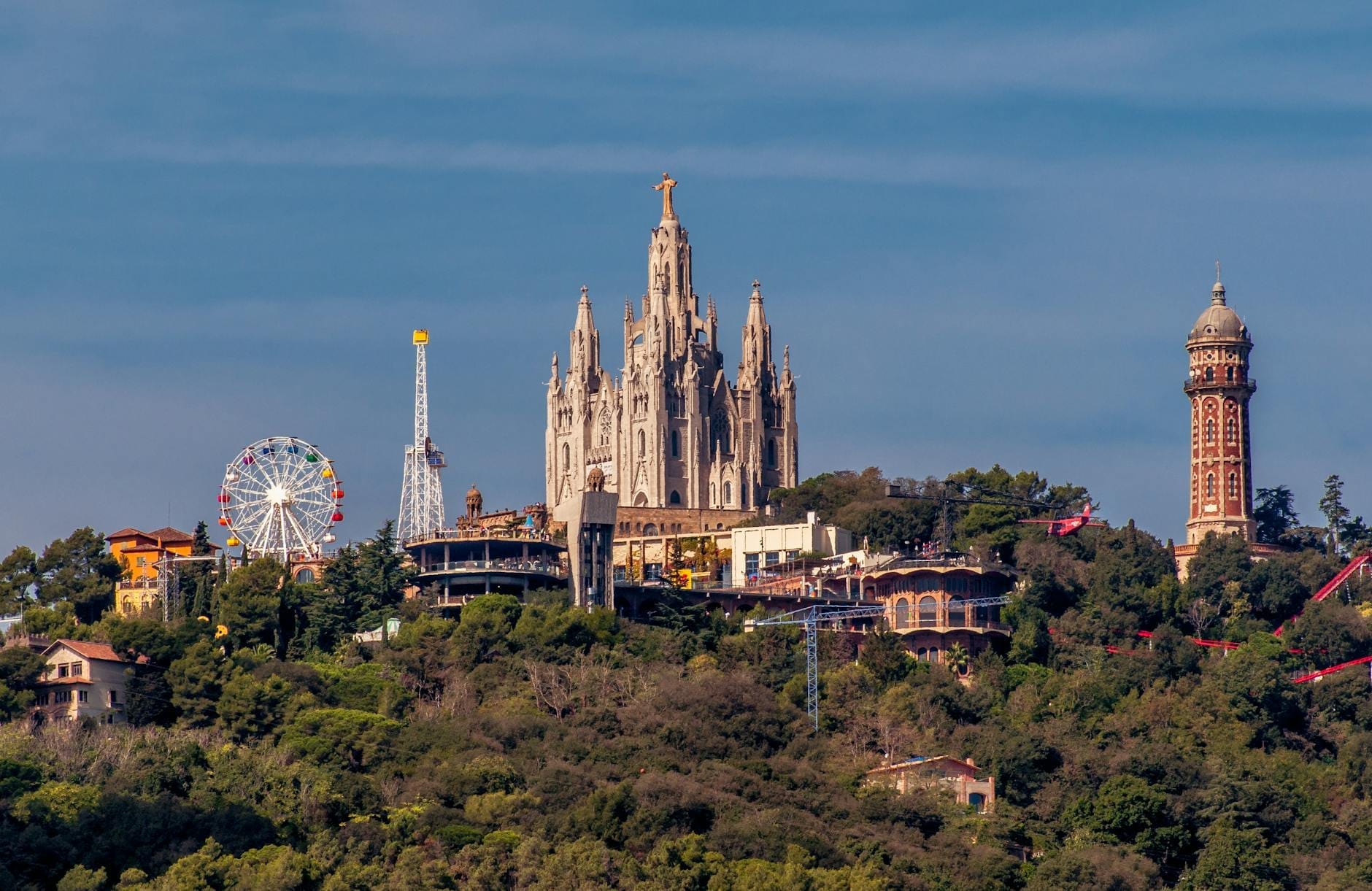 Panoramic view of Tibidabo hill with the Temple Expiatori del Sagrat Cor, vintage amusement park rides, and lush greenery under a clear blue sky.