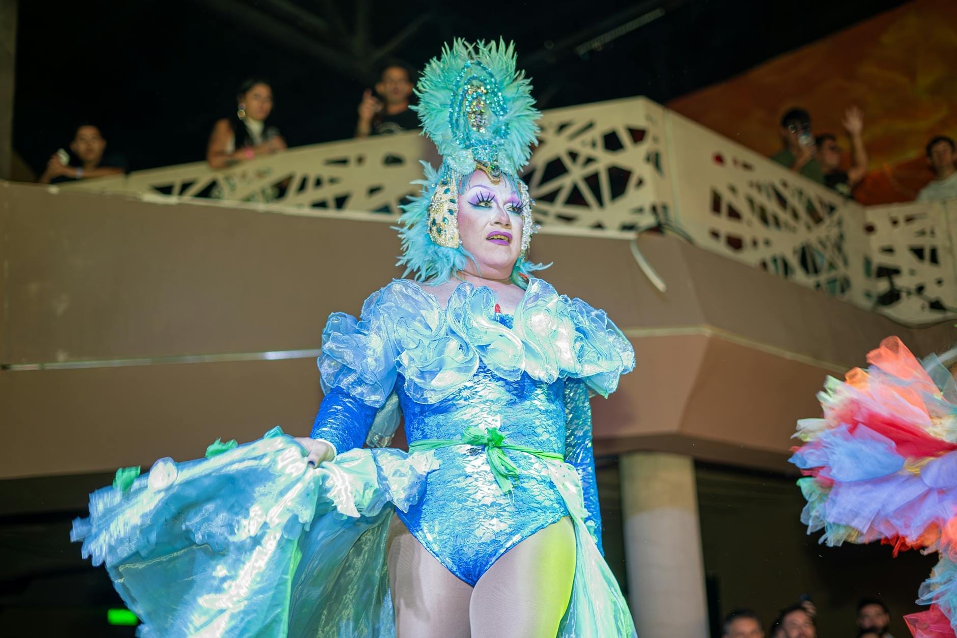 Una vivace artista di cabaret in un elaborato costume con piume blu e paillettes intrattiene il pubblico all'interno di un teatro animato, con spettatori che guardano dai balconi sullo sfondo.