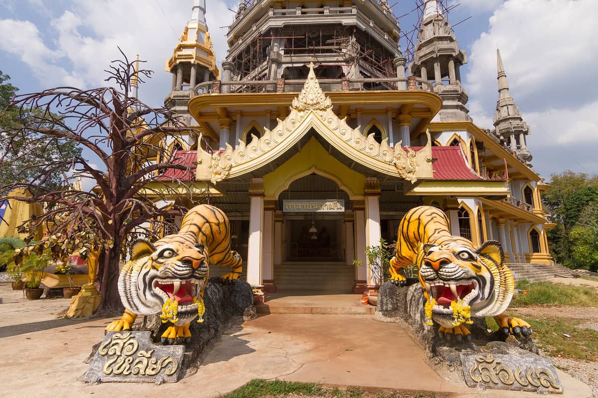 Haupteingang des Tigerhöhlentempels (Wat Tham Suea) in Krabi, Thailand, mit zwei großen Tigerstatuen, die das verzierte Tempelgebäude flankieren, unter einem teils bewölkten Himmel.