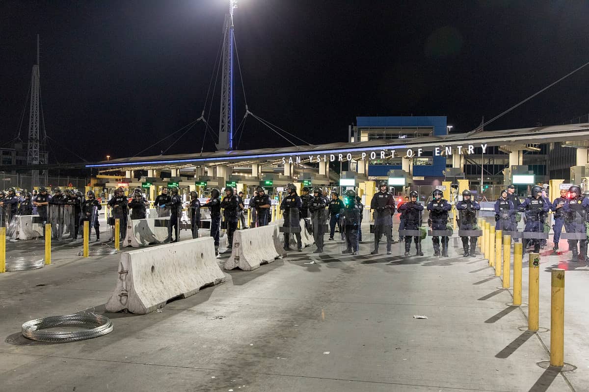 Nächtliche Ansicht des Grenzübergangs San Ysidro mit Reihen von Grenzbeamten in Schutzausrüstung, die unter hellem Licht die Fahrspuren blockieren.