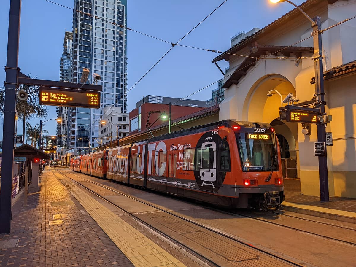 Die Blue Line der San Diego Trolley an der Station Santa Fe Depot, mit einem leuchtend roten Zug, digitalen Anzeigen für San Ysidro und Stadtgebäuden im Hintergrund.