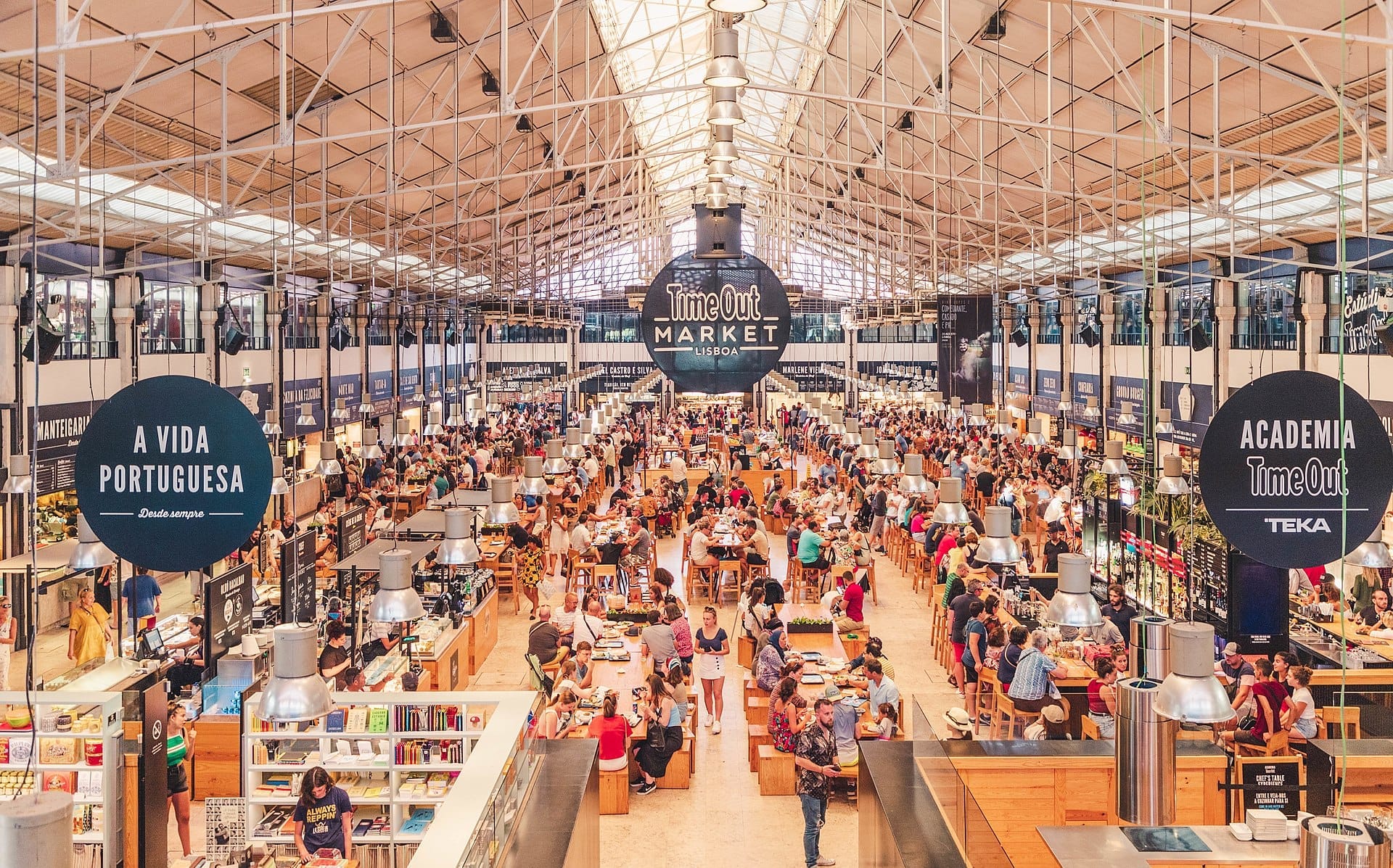 Ampia ripresa degli interni del Time Out Market Lisboa, con folle vivaci, tavoli comuni in legno, bancarelle gastronomiche e soffitto in vetro con struttura in ferro.