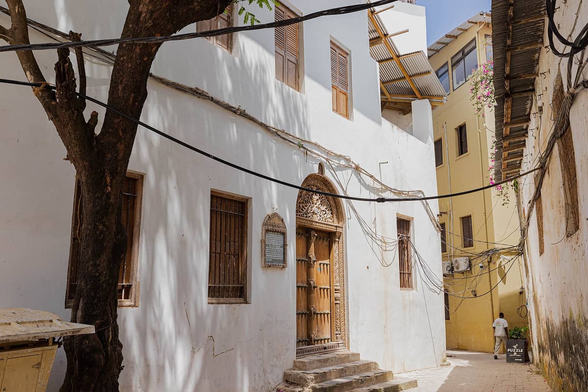 Widok ogólny Domu Tippu Tipa w Stone Town przedstawiający charakterystyczne rzeźbione drewniane drzwi, marmurowe schody, tablicę i kolonialną architekturę budynku w wąskim zaułku.