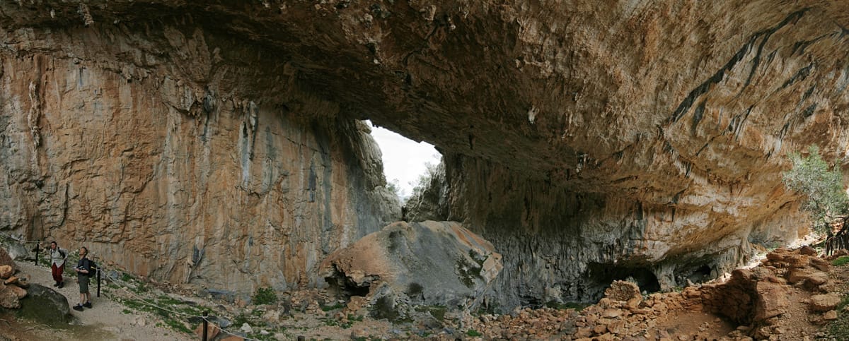 Großer Kalksteinhöhleneingang im nuragischen Dorf Tiscali, mit zwei Wanderern auf einem felsigen Pfad unter den überhängenden Klippen.