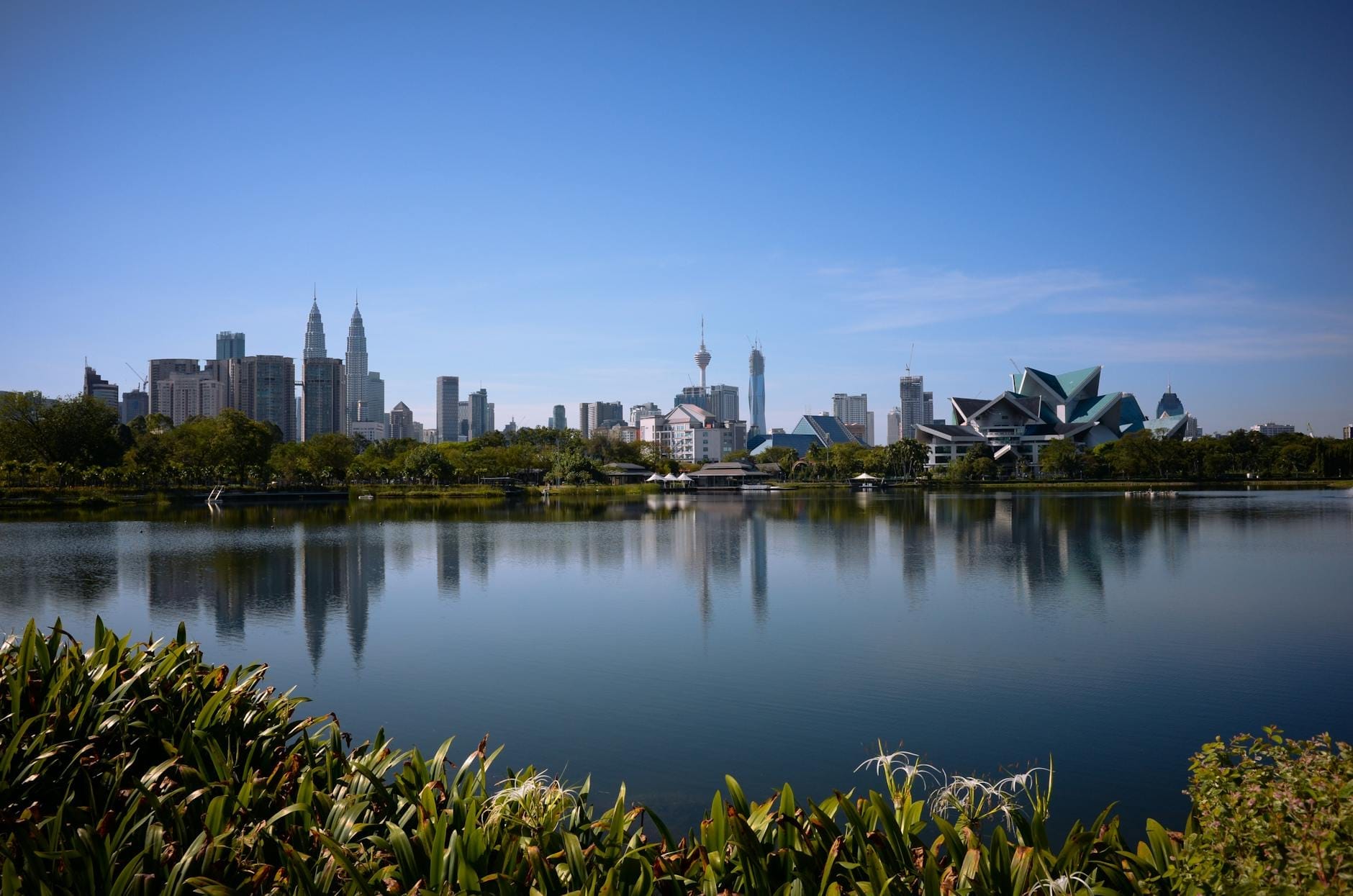 Titiwangsa Park lake with reflections of Kuala Lumpur skyline, including the Petronas Towers and modern architecture, under a clear blue sky.