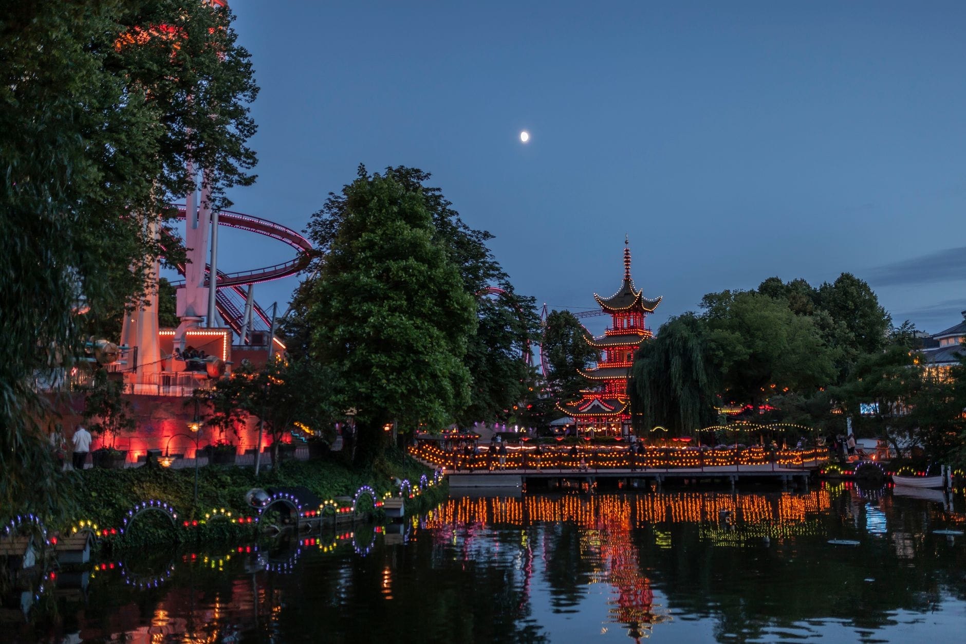 Vista serale di Tivoli Gardens con una pagoda sul lago, lanterne illuminate, alberi e le rotaie di una montagna russa che si riflettono sull'acqua sotto un cielo al crepuscolo.