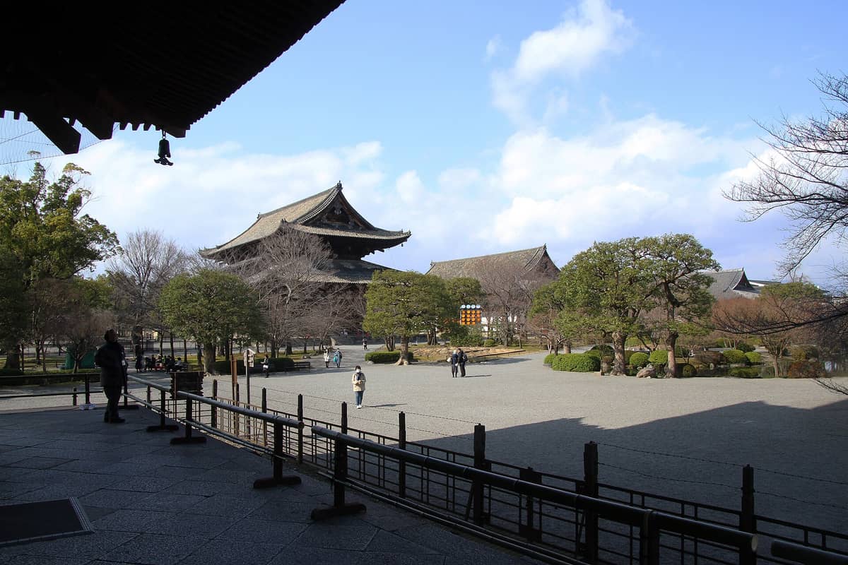 Wide view of Tō-ji Temple in Kyoto showing the main hall with traditional sloping roof, landscaped trees, and open gravel courtyard under a partly cloudy sky.