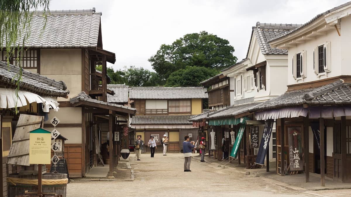 A pedestrian street inside Toei Kyoto Studio Park lined with Edo-period style wooden buildings, signs, and tourists walking in costume and taking photos.
