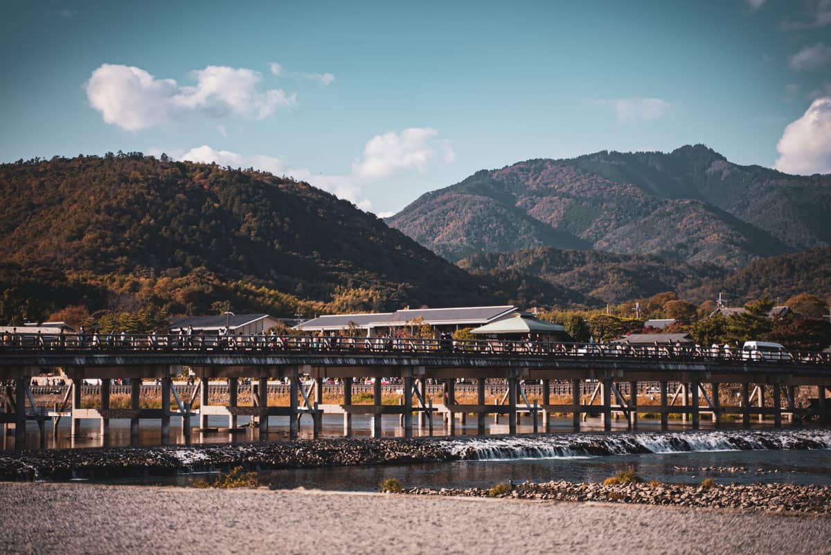 Togetsukyo Bridge stretching over the Oi River with people crossing, backed by forested mountains under a blue sky.