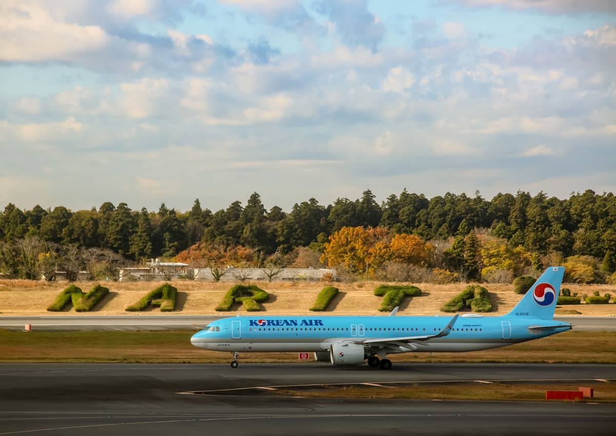 Korean Air plane taxiing at Narita Airport with large greenery spelling 'NARITA' in the background, surrounded by trees and sky.