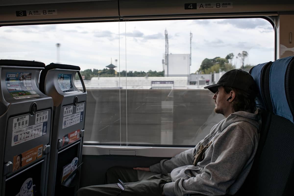 Man gazing out train window in modern Japanese rail car with info panels, headed through suburban Tokyo landscape.