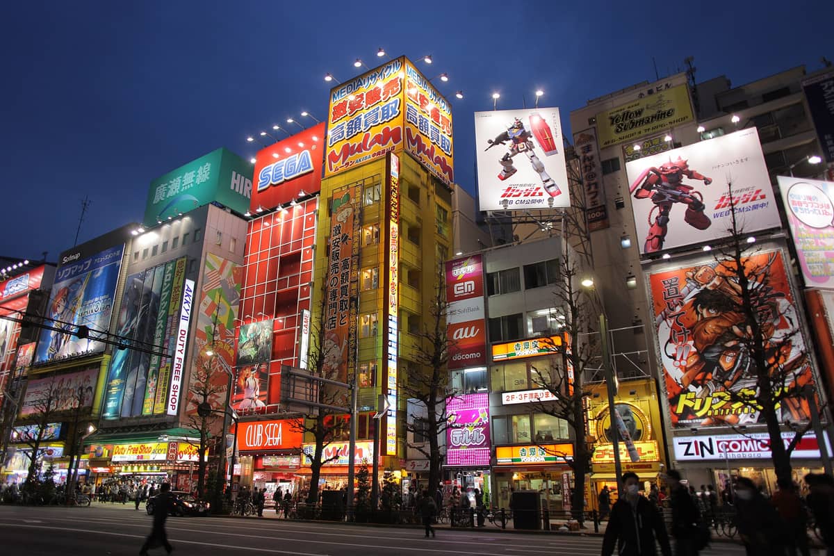 Akihabara street at night with brightly lit anime and game store buildings, giant manga and Gundam billboards, and people walking by.