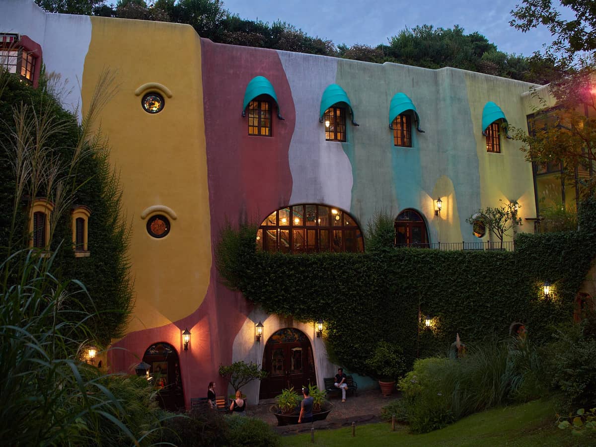 Colorful exterior of Ghibli Museum at dusk, with whimsical architecture, green ivy, arched windows, visitors, and lush garden surroundings.