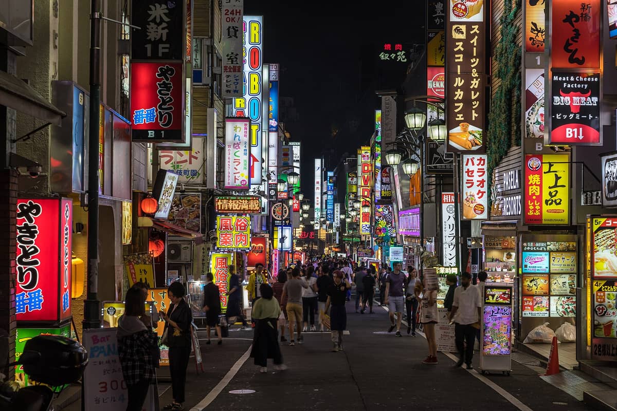 Nighttime street scene in Tokyo’s entertainment district with crowds walking under colorful neon signs and billboards, capturing the city’s vibrant pop culture atmosphere.