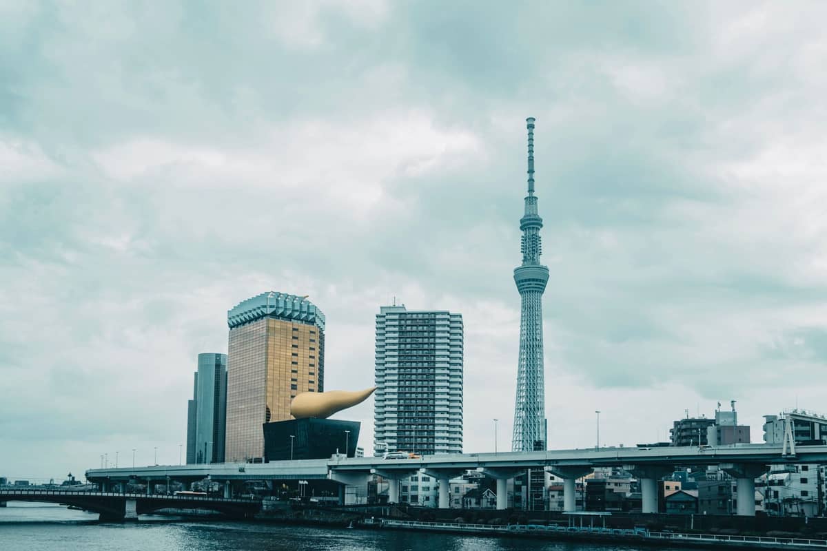 Tokyo Skytree and nearby modern buildings viewed from across the Sumida River under a cloudy sky, showing prominent architectural landmarks.