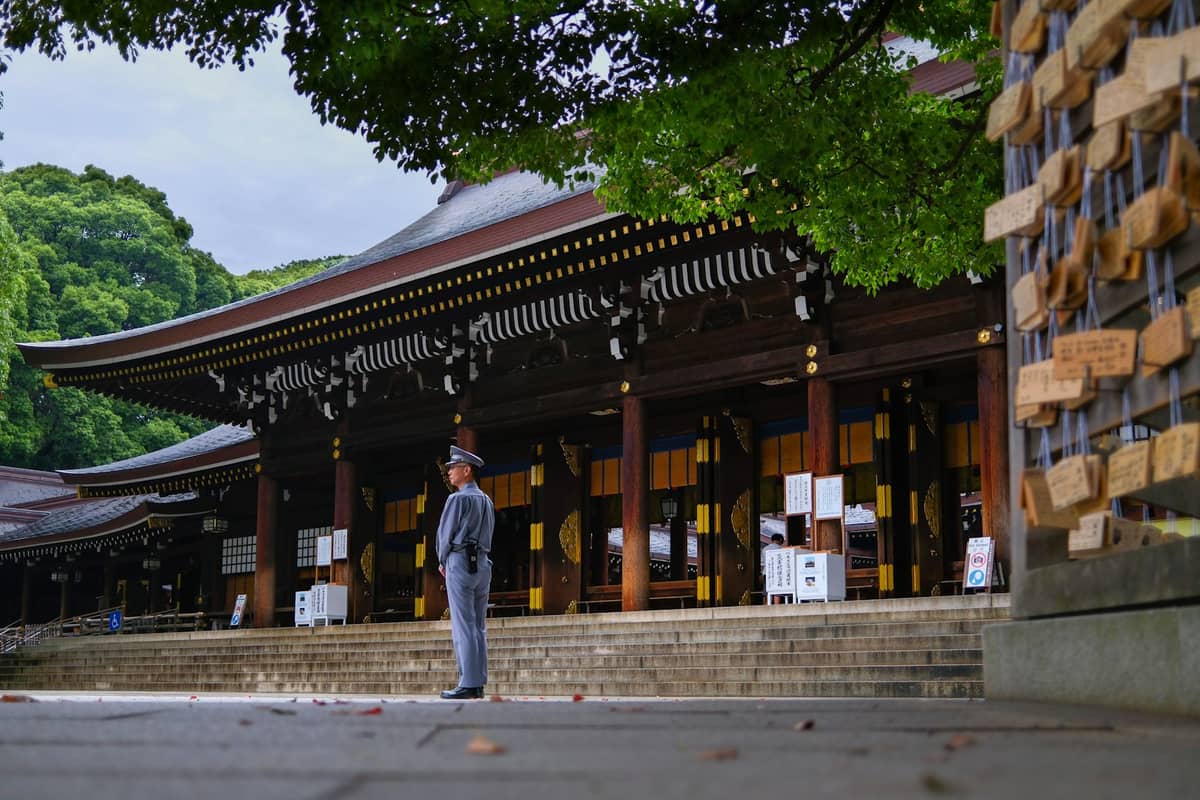 Traditional wooden Shinto shrine building in Tokyo with a uniformed guard, surrounded by trees and wooden prayer plaques in foreground.