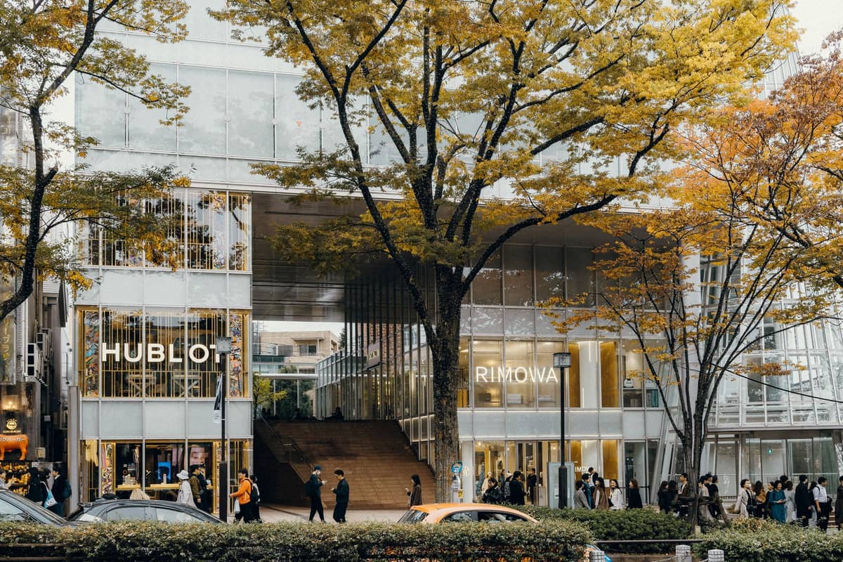 Wide street view of Omotesando in Tokyo with modern glass buildings, upscale shops, and tree-lined sidewalks filled with pedestrians.