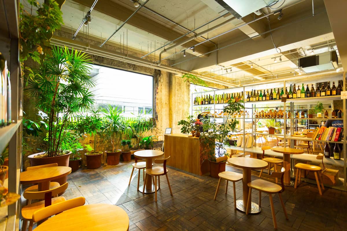 A bright, modern Tokyo café interior with wooden tables, chairs, greenery, books, and sake bottles, capturing the atmosphere of contemporary Japanese café culture.