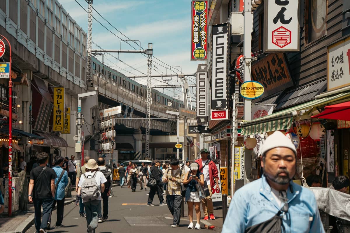 Crowded pedestrian street in Tokyo with restaurant and coffee signs, railway tracks above, and people walking under bright daylight.
