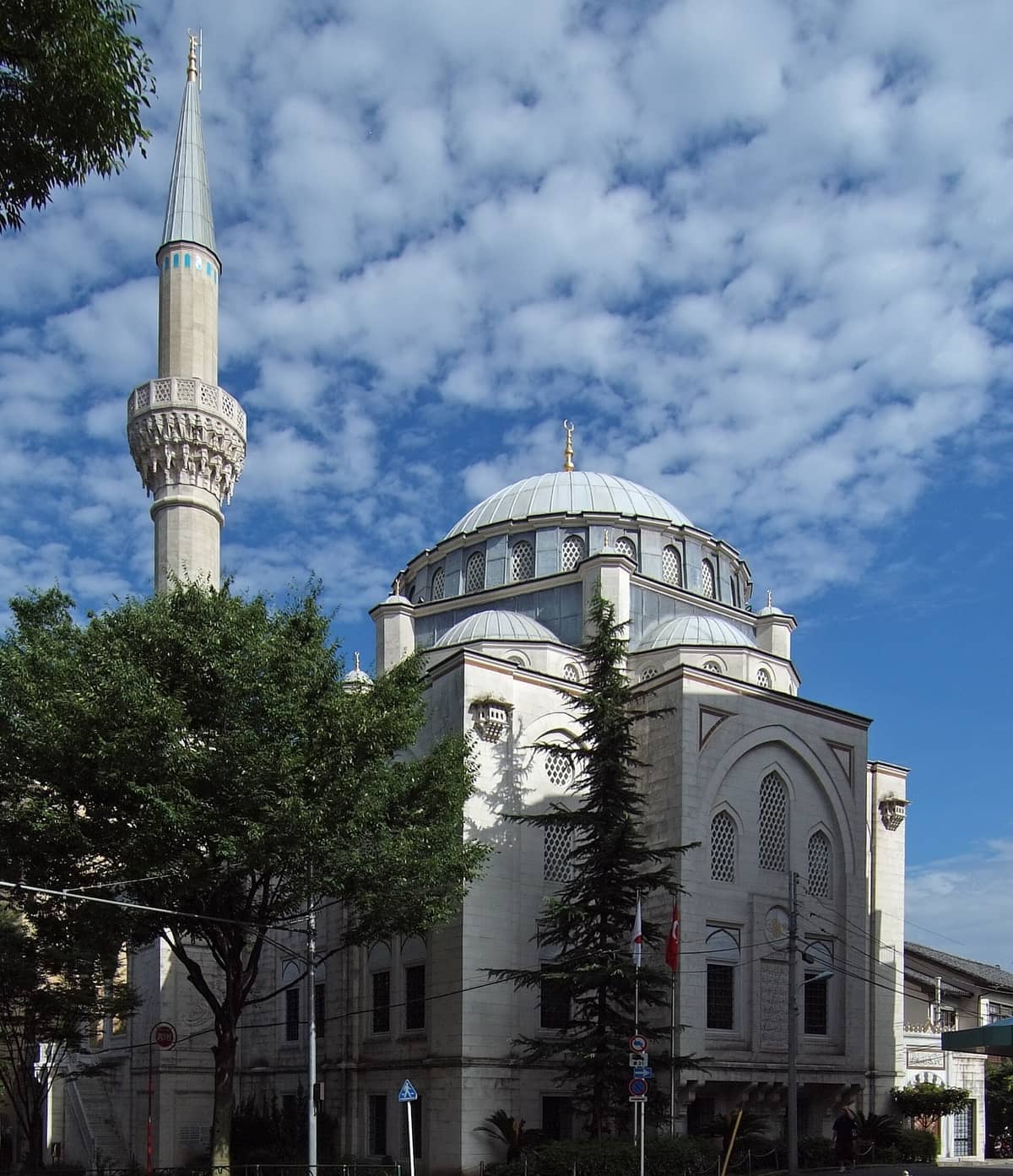 Tokyo Camii mosque with its large central dome, tall minaret, and white Ottoman-style exterior under a partly cloudy sky.