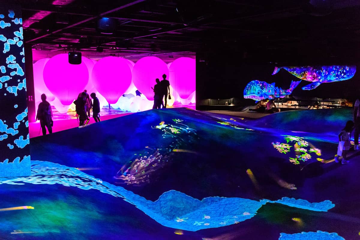 Interior of a Tokyo digital art venue with glowing pink sculptures, people silhouetted, and immersive projected ocean scenes on undulating floors.