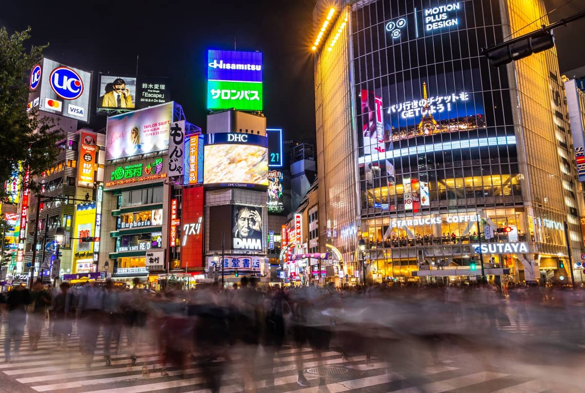Night view of Shibuya Crossing in Tokyo, with bright digital billboards and blurred crowds moving across the famous intersection.