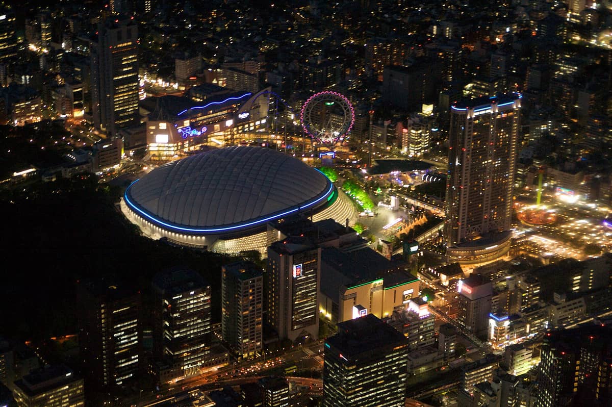 Aerial night view of Tokyo Dome City showing the illuminated Tokyo Dome stadium, amusement park, Ferris wheel, and surrounding city lights.