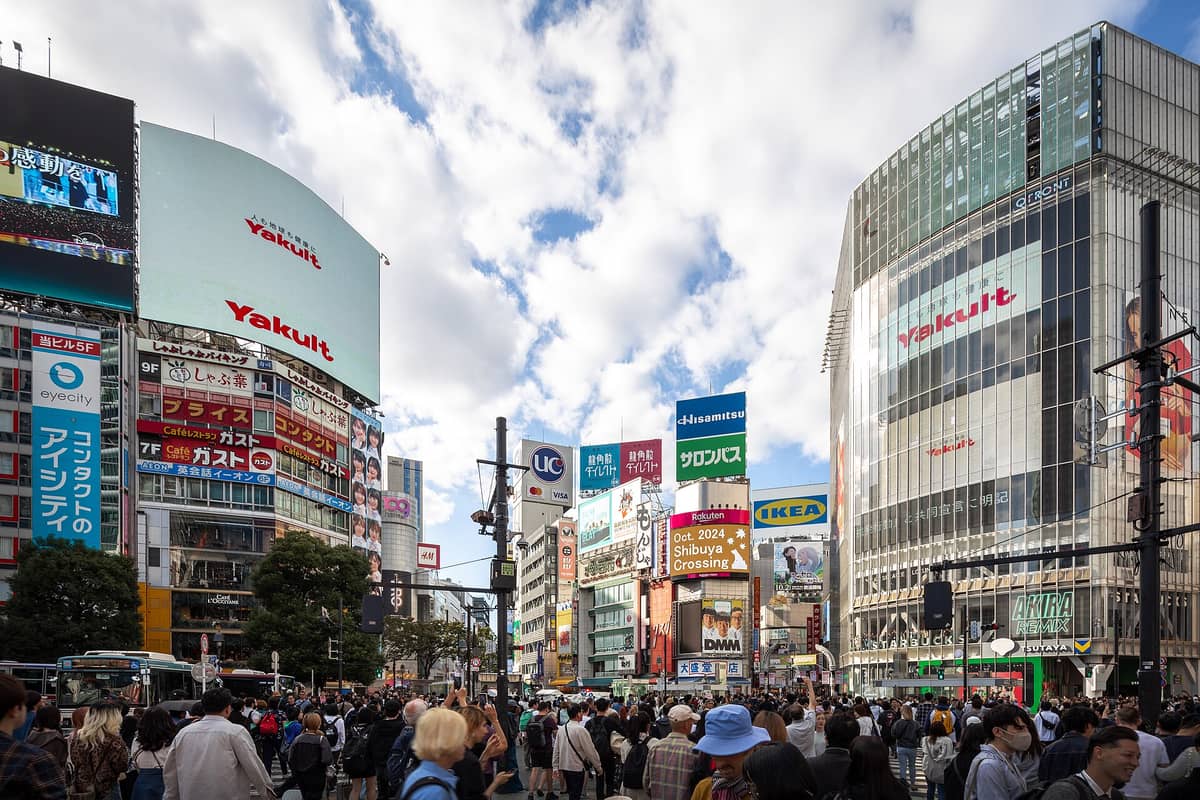 Crowds of people crossing at Shibuya Crossing in Tokyo, surrounded by tall buildings, colorful billboards, and bright daytime sky.