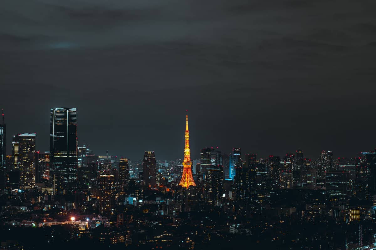 Nighttime cityscape of Tokyo with Tokyo Tower illuminated in orange at the center and modern skyscrapers surrounding it under a dark sky.