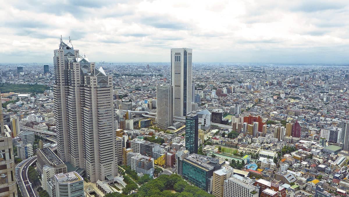 Wide aerial view of central Tokyo with many high-rise buildings, including the distinct Tokyo Metropolitan Government Building, under a cloudy sky.
