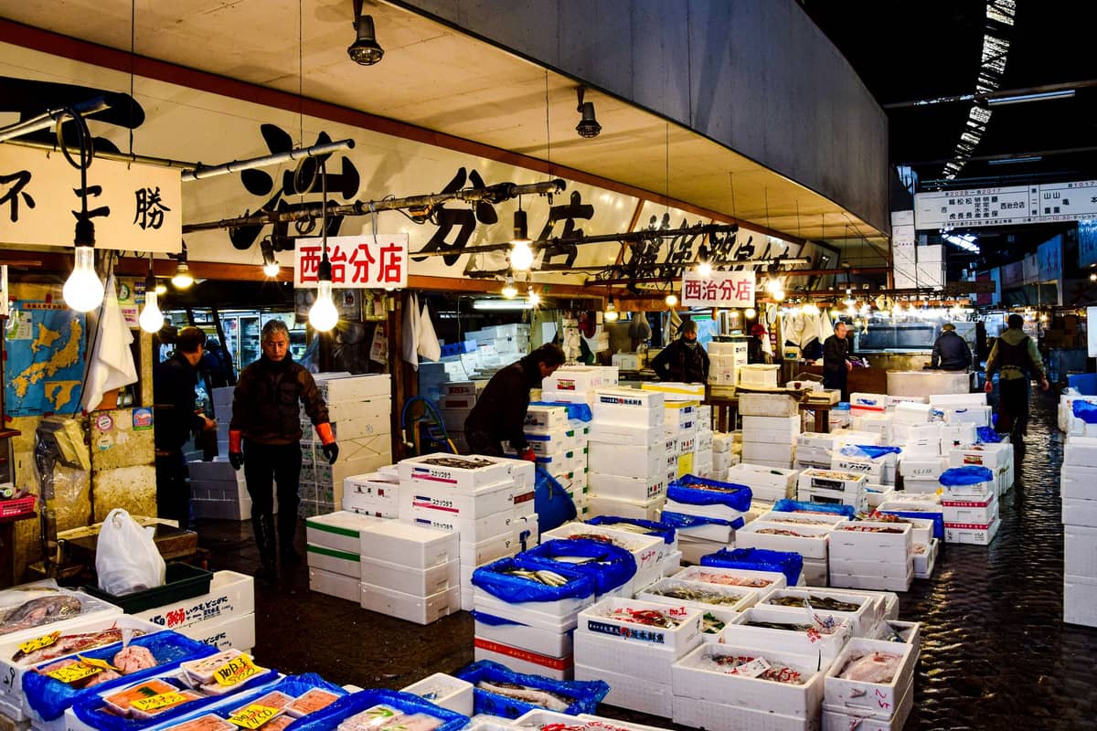 Interior view of the Tsukiji fish market wholesale area with workers, stacks of styrofoam boxes, and Japanese signage above the stalls.