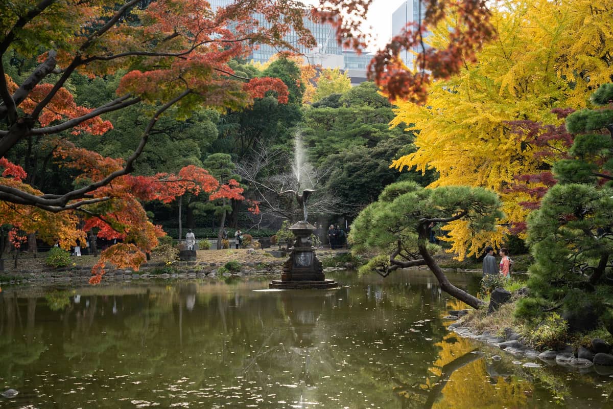 Autumn foliage in Tokyo’s Hibiya Park with vibrant yellow ginkgo and red maple trees framing a pond and fountain, city buildings in the background.