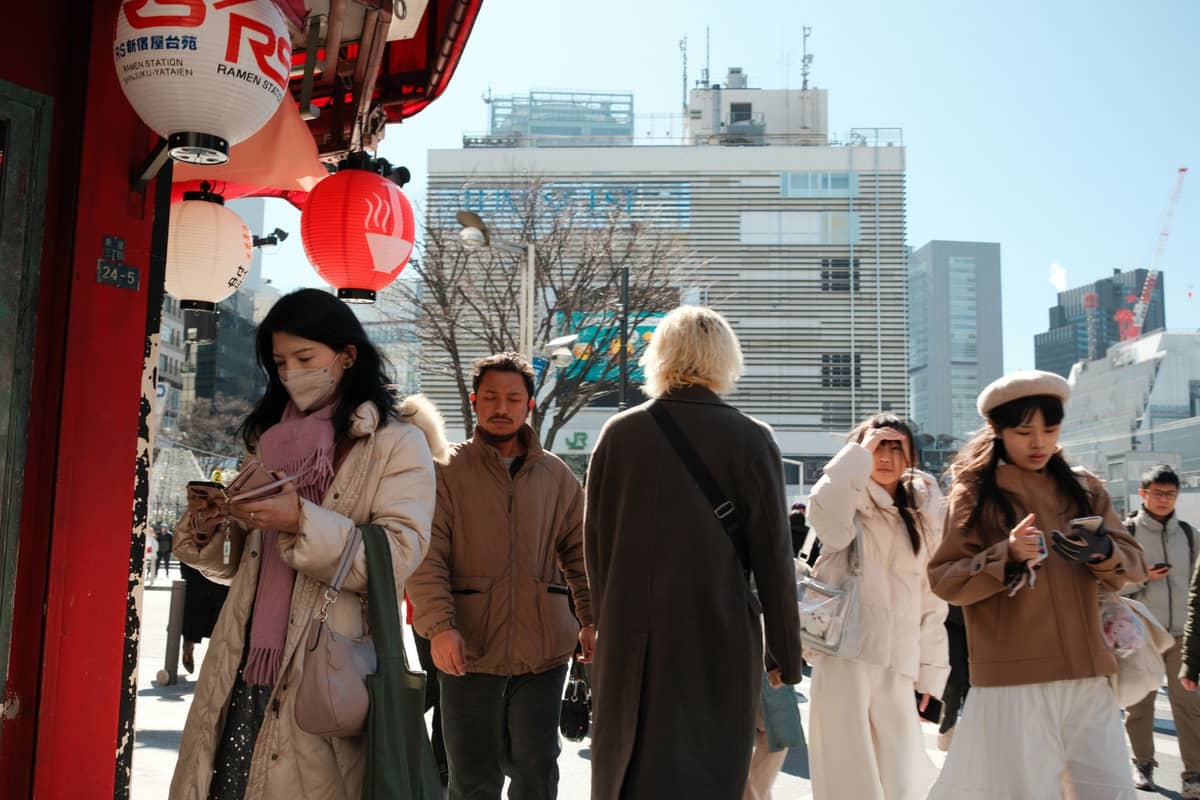 People in winter coats and scarves walk outdoors in urban Tokyo under clear skies, modern buildings in the background, capturing typical December attire and weather.