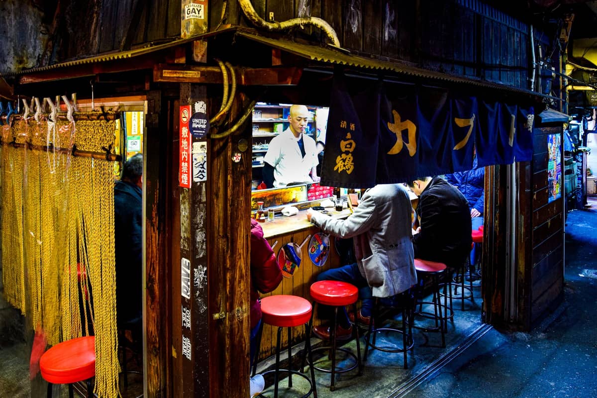 Interior view of a busy Tokyo izakaya with customers sitting at a wooden counter on red stools, chef visible, and traditional decor and noren curtains.