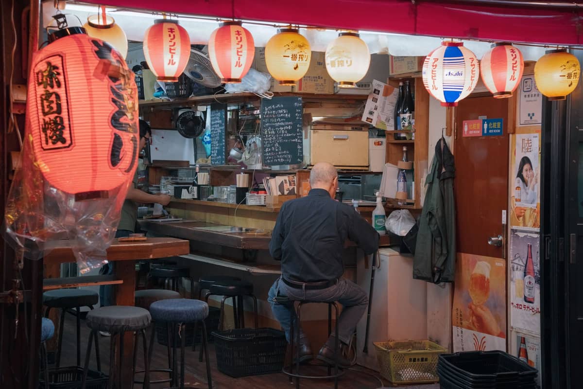 Interior of a small Tokyo izakaya with paper lanterns, a lone customer seated at the counter, and cozy wooden decor.