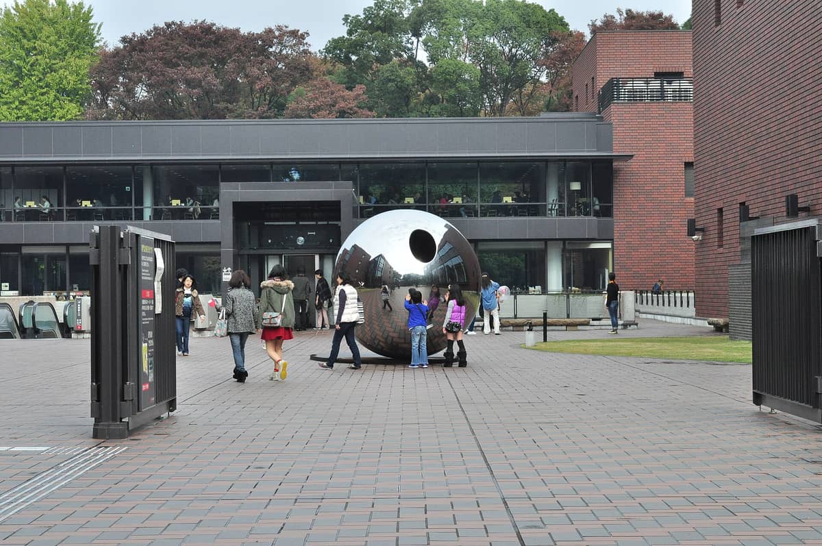 Front plaza of the Tokyo Metropolitan Art Museum with a large mirrored sphere sculpture and visitors walking toward the modern brick building entrance.