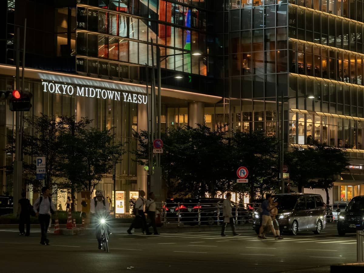 Tokyo Midtown Yaesu’s glass facade glows at night as pedestrians and a cyclist pass by the modern urban complex in Roppongi.