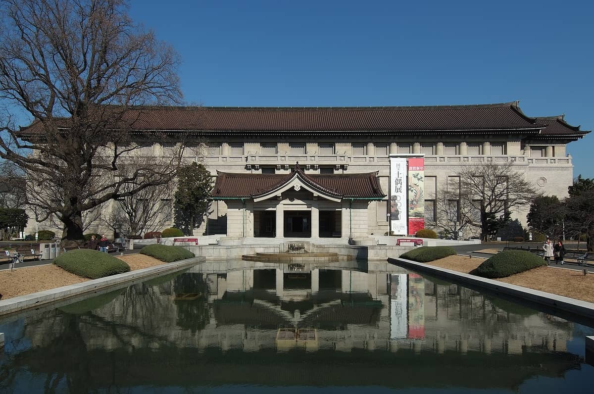 Frontal view of the Tokyo National Museum's main Honkan building with its reflection in the foreground pond under a clear blue sky.