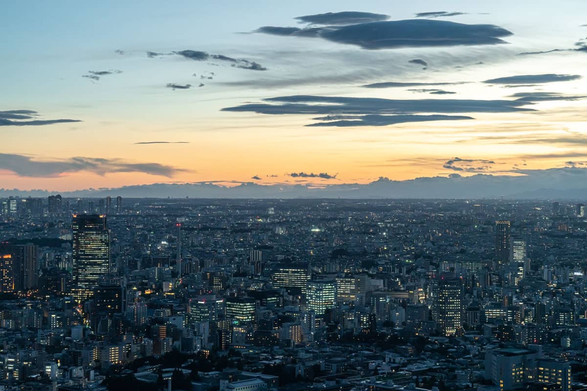 Wide landscape view of Tokyo's dense urban core at sunset, showing sprawling neighborhoods and tall city buildings under a dramatic sky.