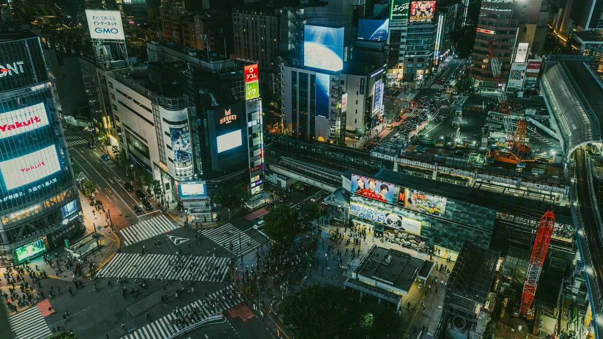 Aerial night view of Shibuya Crossing and surrounding neon-lit buildings in central Tokyo, showing the scale and energy of the nightlife districts.